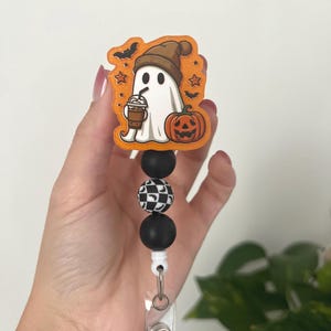Może przedstawiać: Kołowrotek na odznakę o tematyce Halloween z kreskówkowym duchem w brązowej czapce i trzymającym kawę. Duch jest osadzony na pomarańczowym tle z nietoperzami i gwiazdami. Kołowrotek ma czarno-białe szachownice i czarne koraliki.