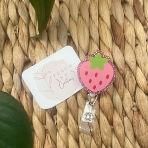 Pu&ograve; includere: Un porta badge a forma di fragola rosa con picciolo verde e semi rossi, caratterizzato da un bordo glitterato. Accanto si trova un cartoncino rettangolare bianco con il testo Peach and Co Creations e un disegno al tratto di un fiore.