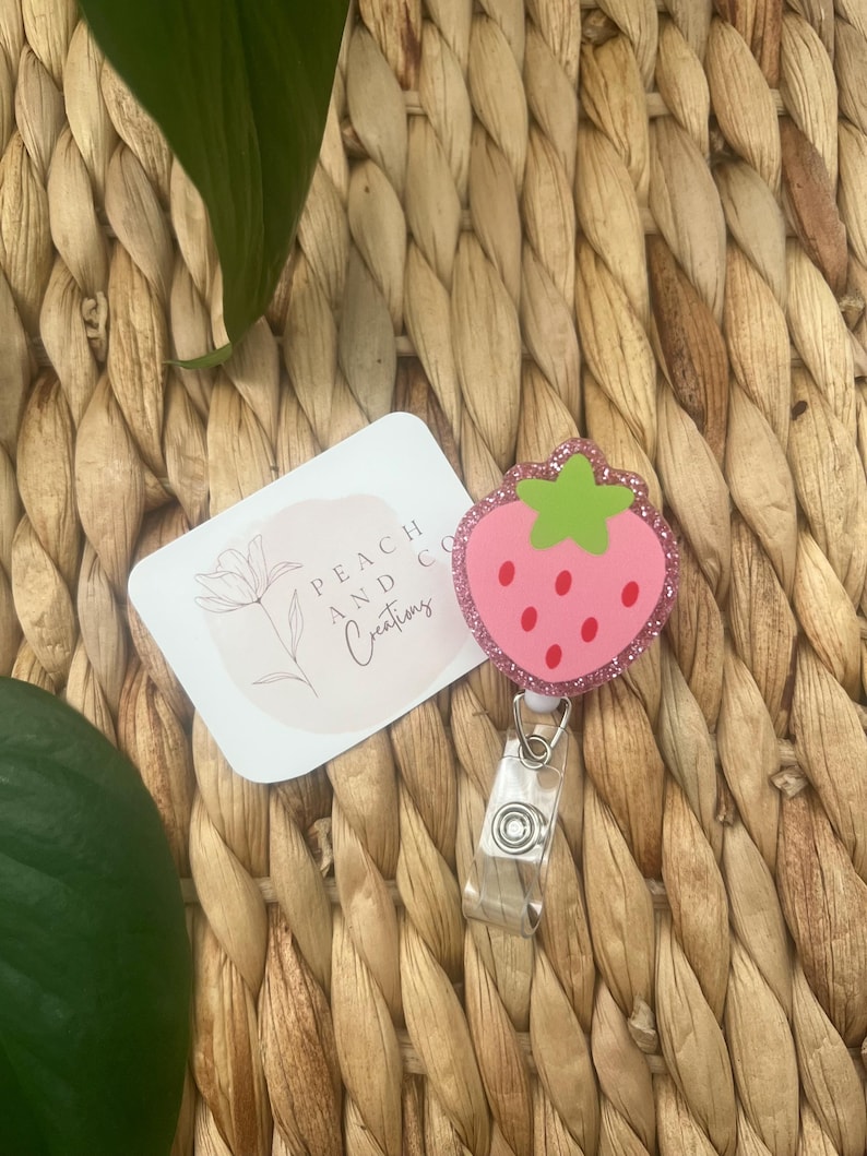Pu&ograve; includere: Un porta badge a forma di fragola rosa con picciolo verde e semi rossi, caratterizzato da un bordo glitterato. Accanto si trova un cartoncino rettangolare bianco con il testo Peach and Co Creations e un disegno al tratto di un fiore.