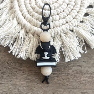 May include: A black bat-shaped keychain with a small book and two speckled beads. The keychain has a black clasp and is set against a macrame coaster and a wooden surface.