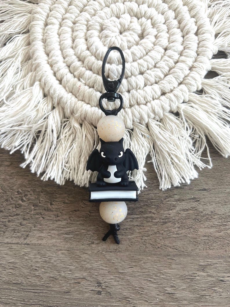 May include: A black bat-shaped keychain with a small book and two speckled beads. The keychain has a black clasp and is set against a macrame coaster and a wooden surface.