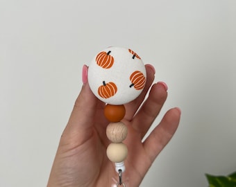 Puede incluir: Un carrete de insignia de tela blanca con un patrón de calabaza. El carrete tiene tres cuentas de madera en colores marrón, beige y crema. El carrete está unido a un soporte de insignia de plástico transparente con un clip de metal.