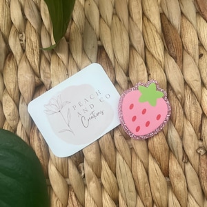 Pu&ograve; includere: Una spilla rosa a forma di fragola con un picciolo verde chiaro e semi rossi, caratterizzata da un bordo glitterato. Accanto si trova un cartoncino rettangolare bianco con il disegno a linee di un fiore e la scritta Peach and Co Creations.