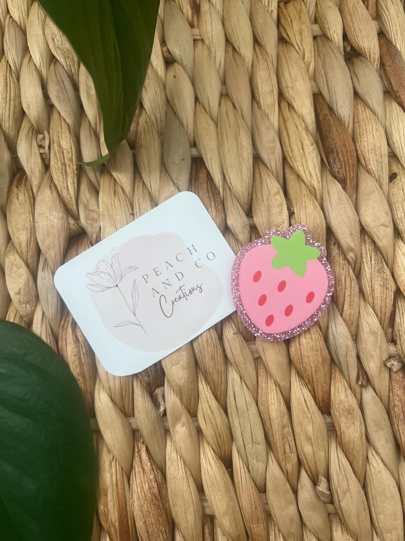 Pu&ograve; includere: Una spilla rosa a forma di fragola con un picciolo verde chiaro e semi rossi, caratterizzata da un bordo glitterato. Accanto si trova un cartoncino rettangolare bianco con il disegno a linee di un fiore e la scritta Peach and Co Creations.