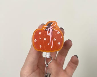 Könnte beinhalten: Ein orangefarbener, kürbisförmiger Ausweishalter mit weißem Blumenmuster und einer rosa Schleife am Stiel. Der Kürbis hat eine glitzernde Oberfläche und ist an einem weißen Kunststoffgehäuse mit einem transparenten Vinylband befestigt.