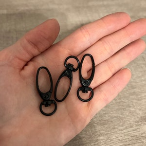 May include: Three black metal swivel clasps with a lobster claw design. Each clasp has a circular ring at the base and a spring-loaded clip. The clasps are shown on a light brown surface.