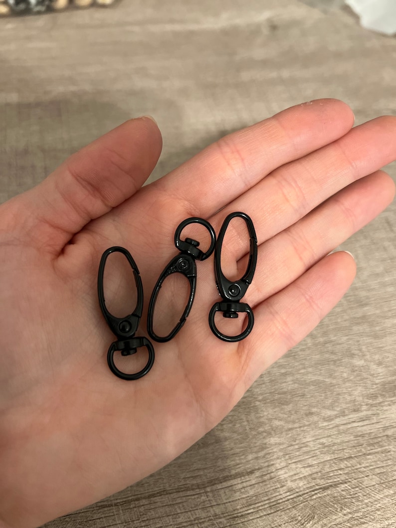 May include: Three black metal swivel clasps with a lobster claw design. Each clasp has a circular ring at the base and a spring-loaded clip. The clasps are shown on a light brown surface.