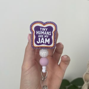 May include: A badge reel featuring a purple glittery toast-shaped topper with the text "TINY HUMANS ARE MY JAM" in white letters. Below the topper are three beads: one white textured bead, one smooth light purple bead, and one small white bead.