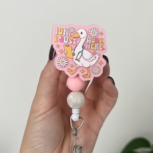 May include: A pink badge reel featuring a cartoon duck with a stethoscope and the text "IDK I JUST WORK HERE". The reel has a clear plastic clip, white cord, and pink and white beads.