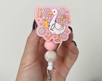 May include: A pink badge reel with the text "IDK I JUST WORK HERE" and a cartoon duck wearing a stethoscope. The reel has a clear plastic clip, white cord, and pink and white beads.