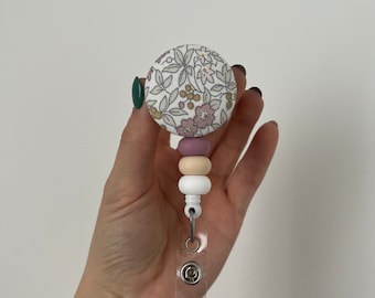 Può includere: Un bottone in tessuto bianco con un motivo floreale, attaccato a un porta badge retrattile con un cinturino in plastica trasparente e una clip argentata. Il porta badge ha una catena di perline rosa, beige e bianca.