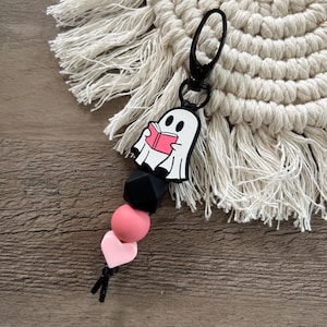 May include: A black and pink silicone keychain with a ghost holding a book charm. The keychain has a black metal clasp.