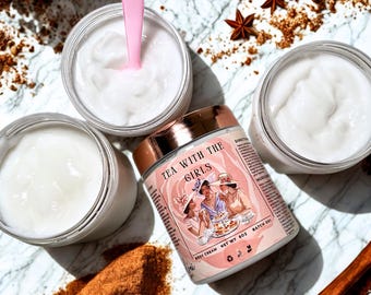 May include: A product shot featuring jars of white body cream, a pink spoon, and a jar labeled "Tea With The Girls" body cream. Cinnamon sticks, star anise, and ground cinnamon are scattered around the jars on a marble surface.