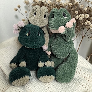 Könnte beinhalten: Drei handgefertigte gehäkelte Dinosaurier-Plüschtiere in Grün- und Beigetönen. Die Dinosaurier haben kleine, runde Augen und sind in einer Gruppe aufgestellt. Die Plüschtiere sind aus weichem, strukturiertem Garn gefertigt, mit rosa Akzenten an Füßen und Armen.