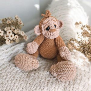 May include: A crocheted brown monkey toy with a sad expression, sitting on a white knitted blanket.