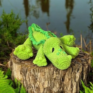 May include: A green crocheted alligator toy sitting on a tree stump in a forest setting. The alligator has a textured, bumpy surface and black eyes. The stump is surrounded by green ferns.