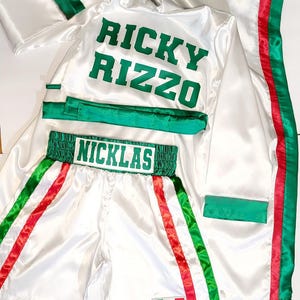 Peut inclure: Ensemble de boxe en satin blanc. La robe est orn&eacute;e de garnitures vertes et rouges, avec "RICKY RIZZO" imprim&eacute; en vert. Le short porte "NICKLAS" sur la ceinture et un &eacute;cusson du drapeau mexicain.