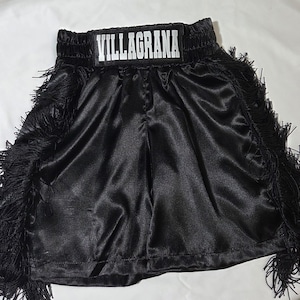 Könnte beinhalten: Schwarze Boxershorts aus Satin mit Fransenbesatz. Der Bund zeigt den Namen "VILLAGRANA" in weißen Blockbuchstaben. Die Shorts haben eine glänzende, glatte Textur und sind für den Sport konzipiert.