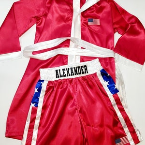 Peut inclure: Ensemble de robe et short de boxe en satin rouge. La robe a une capuche, une bordure blanche et un &eacute;cusson du drapeau am&eacute;ricain. Le short porte le nom "ALEXANDER" &agrave; la taille, avec des &eacute;toiles bleues et blanches et un drapeau am&eacute;ricain.