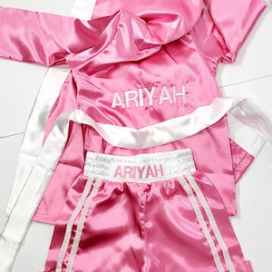 Puede incluir: Conjunto de bata y pantalones cortos de boxeo de satén rosa con ribetes blancos y el nombre "ARIYAH" bordado en ambas piezas.