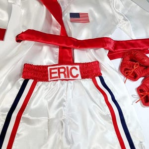 May include: White satin boxing robe with a red sash and an American flag patch. The robe has a hood.  Matching white satin boxing shorts with red trim and blue stripes. The shorts have the name "ERIC" embroidered on the waistband.  Red boxing gloves are included.