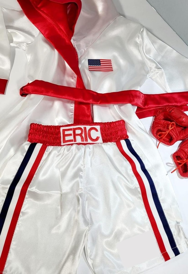 May include: White satin boxing robe with a red sash and an American flag patch. The robe has a hood.  Matching white satin boxing shorts with red trim and blue stripes. The shorts have the name "ERIC" embroidered on the waistband.  Red boxing gloves are included.