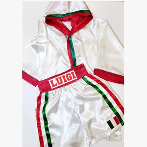Peut inclure: Ensemble de boxe en satin blanc. La robe a une bordure rouge et verte sur la capuche et les manches. Le short porte le nom "LUIGI" en lettres rouges sur la ceinture et des rayures vertes et rouges sur les c&ocirc;t&eacute;s. Un drapeau italien est sur le short.
