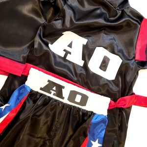Peut inclure: Ensemble de robe et short de boxe noirs. La robe a une capuche avec des accents rouges et bleus, et le short pr&eacute;sente une ceinture blanche avec "AO" en grandes lettres blanches. Le short a des panneaux lat&eacute;raux rouges, blancs et bleus avec une &eacute;toile.