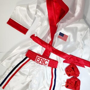 May include: White satin boxing robe with red trim and a red sash. The robe has an American flag patch on the chest. White satin boxing shorts with red and blue trim and the name "ERIC" embroidered on the waistband. Red boxing gloves.