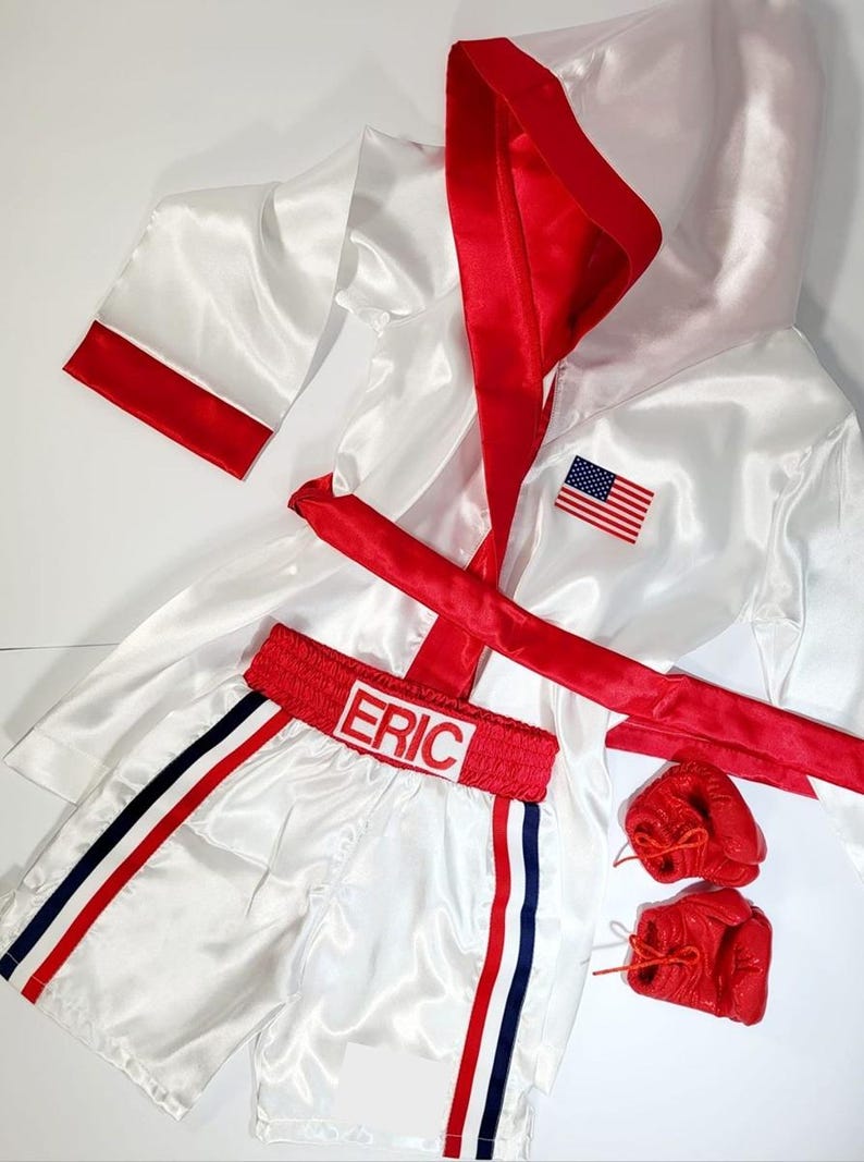 May include: White satin boxing robe with red trim and a red sash. The robe has an American flag patch on the chest. White satin boxing shorts with red and blue trim and the name "ERIC" embroidered on the waistband. Red boxing gloves.