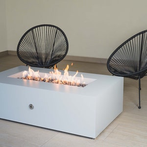 May include: White rectangular fire pit with a flame burning inside. Two black chairs with a woven design are placed on either side of the fire pit.