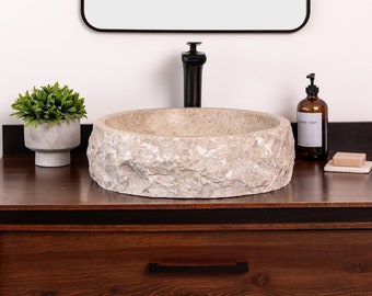 May include: A round, natural stone vessel sink with a rough, textured surface. The sink is mounted on a dark brown wooden vanity with a black drawer pull. A small potted plant and a bottle of hand soap are on the counter.