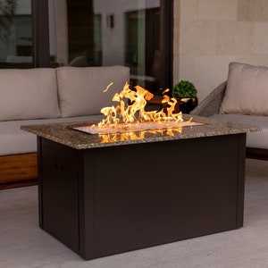 May include: A rectangular fire pit table with a granite top and a dark brown base. The fire pit is lit and has flames rising from the center. The fire pit is surrounded by patio furniture, including a couch and two chairs.