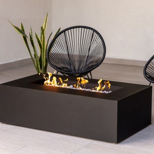 May include: A black rectangular fire pit with flames and white rocks inside. Two black chairs with a woven design are placed on either side of the fire pit.