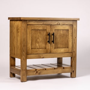 Puede incluir: Un tocador de ba&ntilde;o de madera r&uacute;stica con dos puertas y un estante inferior. El tocador est&aacute; hecho de madera natural con un tinte marr&oacute;n claro. Las puertas tienen manijas de metal negro.
