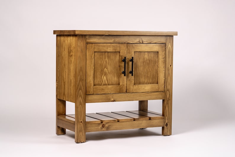 Puede incluir: Un tocador de ba&ntilde;o de madera r&uacute;stica con dos puertas y un estante inferior. El tocador est&aacute; hecho de madera natural con un tinte marr&oacute;n claro. Las puertas tienen manijas de metal negro.
