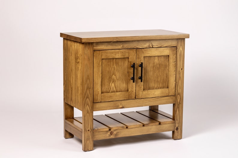 Puede incluir: Un armario de madera con dos puertas y un estante inferior. El armario est&aacute; te&ntilde;ido de un color marr&oacute;n claro y tiene tiradores de metal negro.