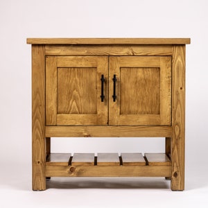 Puede incluir: Un armario de madera con dos puertas y un estante inferior. El armario est&aacute; te&ntilde;ido de un color marr&oacute;n claro y tiene tiradores de metal negro.