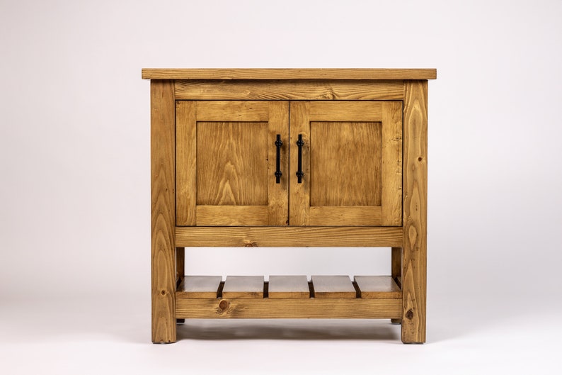 Puede incluir: Un armario de madera con dos puertas y un estante inferior. El armario est&aacute; te&ntilde;ido de un color marr&oacute;n claro y tiene tiradores de metal negro.
