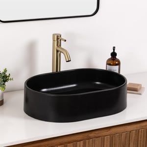 May include: A black oval bathroom sink with a gold faucet. A small plant in a white and brown pot sits to the left. A brown soap dispenser and a bar of soap are on the right. The sink is on a white countertop.