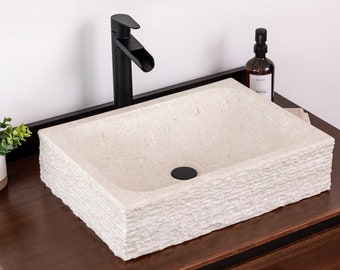 Puede incluir: Un lavabo de piedra blanca con un exterior texturizado y en capas. El lavabo está montado en una encimera de madera oscura con un grifo y un desagüe negros.
