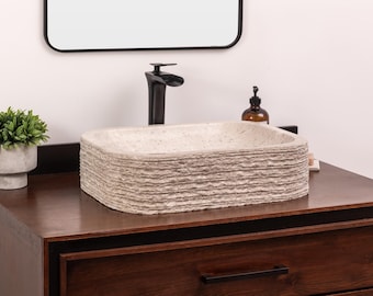 May include: A rectangular, textured, light-colored vessel sink with a stacked design sits atop a dark brown wooden vanity. A black faucet and a small green plant in a concrete pot are also visible. A mirror hangs above.