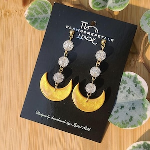May include: A pair of gold crescent moon earrings with dangling crystal beads. The earrings are handmade and feature a unique, textured finish. The card reads "FlawSomePetals" and "Uniquely handmade by Igbol Gill".