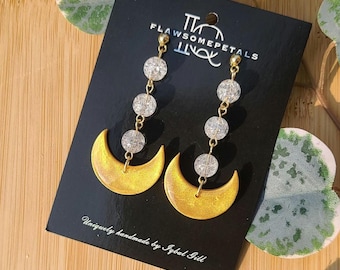 May include: A pair of gold crescent moon earrings with dangling crystal beads. The earrings are handmade and feature a unique, textured finish. The card reads "FlawSomePetals" and "Uniquely handmade by Igbol Gill".