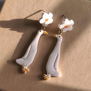 May include: A pair of white ceramic earrings with a small flower charm and a goose design. The flower charm has a yellow centre.