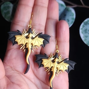 May include: A pair of gold and black dragon earrings. The dragons are in flight with their wings spread. The earrings are made of polymer clay and have gold hooks.