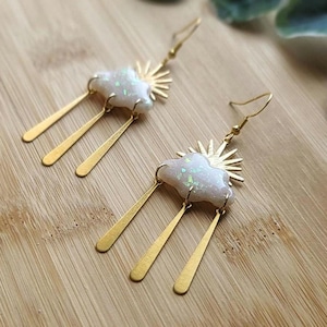 Peut inclure: Une paire de boucles d'oreilles en or avec un design de nuage et de soleil blanc. Les nuages sont en opale irisée et le soleil a un design en étoile. Les boucles d'oreilles ont trois barres dorées pendantes.