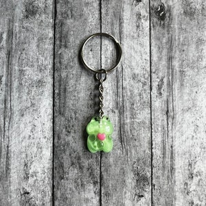 May include: A green gummy bear keychain with a pink heart on its chest. The keychain is attached to a silver chain and ring.