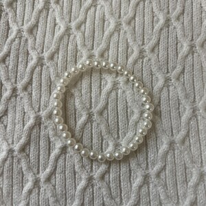 May include: A white pearl bracelet resting on a white, textured surface. The bracelet is composed of numerous small, round, white pearls strung together to form a complete circle. The background is a white, knitted fabric.