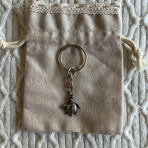 May include: A silver-colored turtle charm keychain hanging from a metal ring, presented with a beige drawstring pouch. The pouch is trimmed with lace and rests on a textured, off-white surface.